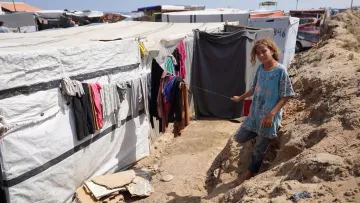 L'image montre un camp de tentes ou de structures temporaires. À gauche, des vêtements sont accrochés à une corde à linge, tandis qu'une jeune fille se trouve au premier plan, tenant une sorte de bâton. Elle porte une tunique et semble se tenir sur un chemin étroit entre les tentes. Le décor est marqué par des bords de sol en terre et la structure du camp qui se développe à l'arrière-plan. L'atmosphère semble celle d'un environnement difficile, lié à des conditions de vie précaires.