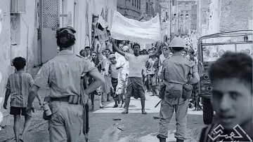 The image depicts a historical scene in a narrow street, likely during a protest or demonstration. In the foreground, a group of people is visible, with a man raising a banner or flag, indicating a spirited display. There are several individuals, including children, observing the scene. Two uniformed officers can be seen in the foreground, suggesting a presence of law enforcement. The atmosphere appears tense, with people engaged in an active moment, highlighting social or political unrest. The black-and-white format emphasizes the historical context of the image.