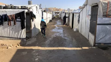 L'image montre un camp de réfugiés, avec des tentes blanches et des structures temporaires alignées des deux côtés d'un chemin étroit. Le sol semble boueux, probablement à cause de la pluie. On peut voir des vêtements accrochés à l'extérieur des tentes. Une personne, vêtue de manière simple, marche le long du chemin central. L'environnement semble rudimentaire et surpeuplé, avec des signes de vie quotidienne.