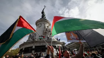 L'image montre une scène de manifestation avec des participants brandissant des drapeaux palestiniens. En arrière-plan, on peut voir un monument, probablement une statue, qui est entouré d'un ciel nuageux. Les personnes présentes semblent exprimant des sentiments forts, et l'atmosphère générale est celle d'un rassemblement public engagé et solennel.