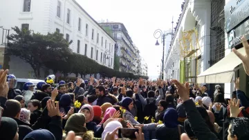 The image depicts a large crowd of people gathered in an urban setting, possibly during a protest or demonstration. Many individuals are raising their hands, and a sense of unity and collective action is present. The atmosphere appears to be lively, and the crowd is surrounded by buildings, suggesting a city environment. Some police presence is visible, indicating that the gathering may be significant or organized and possibly involves a call for change or social issues. The crowd includes diverse individuals, wearing a range of clothing styles, with some wearing headscarves.