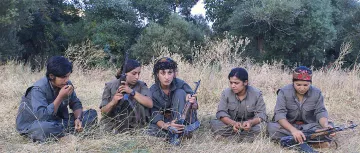 L'image montre un groupe de cinq femmes assises dans une prairie, entourées de végétation. Elles portent des vêtements militaires et semblent concentrées sur une activité commune. Certaines d'entre elles tiennent des armes à feu, tandis que d'autres ont les mains posées sur leurs genoux. Leurs expressions faciales et leur posture suggèrent une ambiance sérieuse et déterminée.