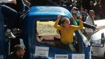 L'image montre un groupe de personnes rassemblées autour d'un camion. Une femme, avec les bras levés, semble exprimer sa frustration ou son indignation. À côté d'elle, un enfant tient une pancarte avec l'inscription "Système Dégage". Le camion est décoré de quelques autres affiches, et la scène se déroule probablement lors d'une manifestation ou d'une protestation. L'atmosphère semble énergique et engagée, avec d'autres personnes en arrière-plan qui participent à la même dynamique.