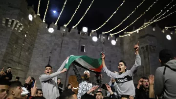 L'immagine mostra un gruppo di persone riunite in un contesto festivo, probabilmente in un evento pubblico. I partecipanti stanno sventolando una bandiera, che sembra essere quella palestinese, e sono visibilmente entusiasti. Sullo sfondo, si può notare una struttura medievale, che suggerisce un'atmosfera di celebrazione. Le luci appese creano un'atmosfera suggestiva e notturna, mentre i volti delle persone esprimono gioia e partecipazione.