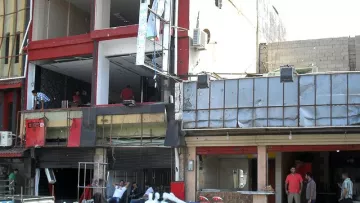 L'image montre un environnement urbain avec des bâtiments partiellement en ruines. On peut voir des façades de bâtiments endommagées, des débris éparpillés au sol et des personnes dans les environs, certaines visiblement en train de travailler sur la reconstruction. Il y a également des voitures garées à proximité. L'atmosphère semble être celle d'une zone en cours de rénovation ou de nettoyage après des dommages.