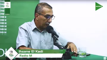 L'image montre un homme assis derrière un micro dans un studio de radio. Il porte une chemise à manches courtes et des lunettes. Sur la table devant lui, il y a un verre d'eau et un téléphone. L'arrière-plan est vert avec des éléments de branding pour "Radio M". Le nom "Ihsane El Kadi" est affiché en bas de l'image.