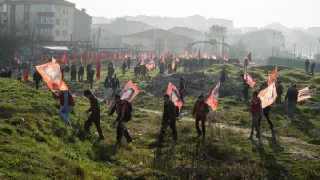 L'image montre un rassemblement de personnes marchant sur un terrain, probablement lors d'une manifestation ou d'une célébration. Les participants portent des drapeaux rouges et des bannières avec des portraits, suggérant un soutien à une cause ou à un leader spécifique. En arrière-plan, on peut voir un paysage urbain flou avec des bâtiments. L'atmosphère semble animée, et la luminosité indique qu'il pourrait s'agir d'une matinée ou d'un après-midi. La présence de nombreux drapeaux rouges donne une impression de rassemblement massif.