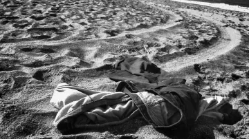 L'image montre un paysage de plage en noir et blanc, où des vêtements abandonnés sont éparpillés sur le sable. On peut voir des traces laissées par les pas dans le sable, créant un motif dans la texture. À l'arrière-plan, la mer se déroule tranquillement, et le ciel est clair. L'ensemble dégage une atmosphère de solitude et d'abandon.