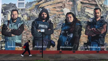 L'immagine mostra un murale che ritrae quattro giornalisti con abbigliamento di protezione e giubbotti con la scritta "PRESS". Sono posizionati in modo prominente davanti a un contesto di distruzione, simile a un'area colpita da un terremoto. Una persona sta camminando lungo il marciapiede di fronte al murale. I volti dei giornalisti esprimono determinazione e serietà, sottolineando il loro lavoro e l'importanza della libertà di stampa.