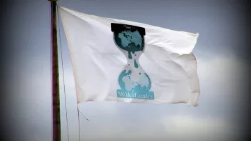 L'image montre un drapeau blanc flottant en l'air, sur lequel est représenté le logo de WikiLeaks. Ce logo se compose d'une silhouette d'une horloge de sable, avec des gouttes tombant d'elle, et une carte du monde en arrière-plan. Le fond apparaît nuageux, ce qui ajoute une ambiance dramatique à l'image.