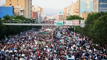 L'immagine mostra una grande folla di manifestanti in una strada cittadina. Le persone sembrano riunite per un evento significativo, con molti che portano cartelli e striscioni. I palazzi al bordo della strada sono visibili, così come alcune automobili parcheggiate. La scena trasmette una forte sensazione di unione e mobilitazione collettiva. Le strade sono affollate e l'atmosfera è intensa, suggerendo che si tratta di una protesta o di una manifestazione pubblica.