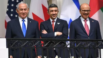 L'image montre trois hommes debout sur un balcon, avec des drapeaux en arrière-plan, dont ceux des États-Unis, du Canada, et d'Israël. Les hommes semblent souriants et détendus, portant des costumes formels. Il y a des fleurs blanches qui décorent le balcon. La scène exprime une ambiance de diplomatie ou de réunion internationale.