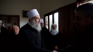 Group of men, some with white hats and beards, shaking hands and wearing dark clothing in a room with windows.