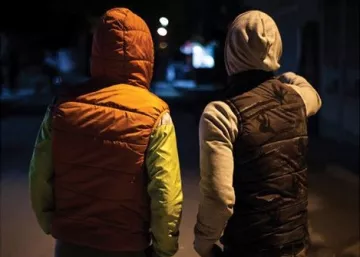 L'image montre deux personnes de dos, se tenant à proximité l'une de l'autre. L'une porte un gilet d'une couleur orange avec des manches vertes, tandis que l'autre est vêtu d'un gilet noir et d'une capuche grise. Ils semblent se trouver dans une rue sombre, avec quelques lumières au loin, ce qui donne une ambiance nocturne à la scène. L'une des personnes pointe quelque chose du doigt, suggérant une conversation ou une observation partagée.