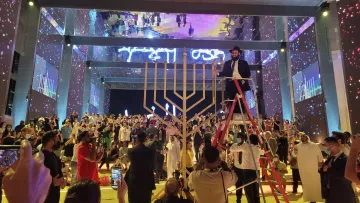 L'image montre une scène festive dans une grande salle. Au centre, une grande menorah (un candélabre à sept branches) est mise en avant. Un homme en costume est sur une échelle, probablement en train d'allumer une bougie. La salle est remplie de personnes, certaines prenant des photos, tandis que d'autres regardent attentivement. Les murs sont décorés avec des éclairages colorés et des projections, créant une atmosphère vibrante et festive. L'ensemble de l'événement semble célébrer une occasion spéciale, probablement liée à la culture ou à la tradition juive.