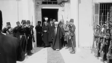 L'image montre un groupe de personnes en costume traditionnel, saluant respectueusement un homme au centre qui semble être une figure de proue. L'ambiance est formelle et cérémonieuse. On peut voir des soldats en uniforme tenant des armes et des dignitaires en robes longues. L'architecture en arrière-plan indique que la scène se déroule probablement dans un bâtiment important ou historique. Les expressions des personnes présentent témoignent d'un moment solennel ou d'une cérémonie officielle.