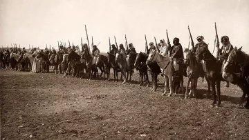 The image depicts a line of mounted warriors or soldiers, likely from a historical context. They are on horseback, holding spears or similar weapons, and are dressed in traditional clothing that suggests their cultural identity. The setting appears to be an open, dry landscape, possibly a desert or arid region. The overall sepia tone gives the photograph an aged appearance, typical of early 20th-century photography. The formation and attire indicate that it could represent a military or ceremonial gathering.