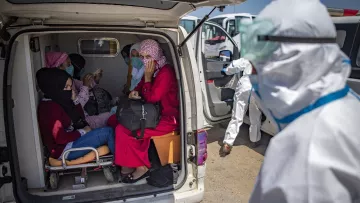 L'image montre un groupe de personnes à l'intérieur d'une ambulance. On peut voir plusieurs femmes assises, certaines portant des masques et des vêtements colorés. Une femme porte un hijab et semble discuter avec les autres. À l'extérieur, une personne en tenue de protection, probablement un professionnel de la santé, s'approche. L'environnement semble être une zone d'intervention médicale. La scène évoque une atmosphère de soin et d'assistance dans un contexte de santé.