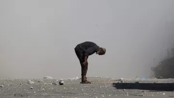 A man bends over on a dusty street, surrounded by debris and smoke.
