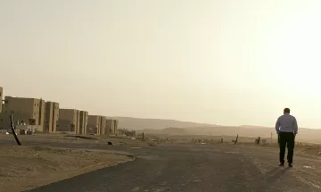 L'image montre un homme marchant seul sur une route déserte. On peut voir en arrière-plan des bâtiments modernes qui semblent inachevés ou abandonnés, avec un paysage désertique s'étendant à l'horizon. La lumière du soleil semble douce, suggérant une ambiance calme et un peu mélancolique. Le ciel est clair, et l'ensemble de la scène évoque une sensation de solitude et d'isolement.