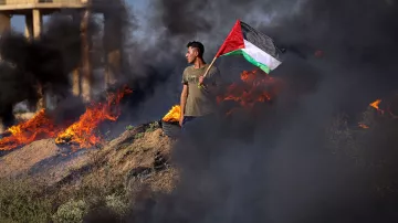 L'image montre une personne debout sur un terrain où des feux brûlent, produisant une épaisse fumée noire. Cette personne tient fièrement un drapeau palestinien, qui est composé de bandes rouge, noire, blanche et verte. Le paysage semble chaotique en raison des flammes entourant le manifestant, créant une atmosphère intense. Les éléments visuels de l'image évoquent des sentiments de militantisme et de détermination.