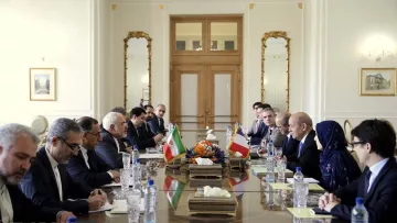 L'image montre une salle de réunion avec une grande table au centre. De chaque côté de la table, plusieurs personnes sont assises, participent à une discussion. Les drapeaux de l'Iran et d'un autre pays sont visibles sur la table. L'environnement est élégant, avec des décorations raffinées et de grandes fenêtres. On peut aussi voir des fleurs au centre de la table. Les participants semblent concentrés et engagés dans la conversation.