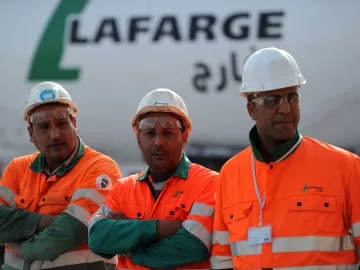 L'image montre trois ouvriers portant des vêtements de sécurité orange et des casques blancs. Ils se tiennent debout avec les bras croisés, affichant une attitude sérieuse. En arrière-plan, on peut voir un camion ou un véhicule avec le logo de Lafarge, une entreprise connue dans le secteur des matériaux de construction. L'environnement semble être un chantier ou une zone industrielle.