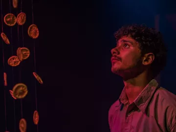 L'immagine mostra un giovane uomo che osserva con meraviglia degli agrumi essiccati appesi in aria. La sua espressione è attenta e sognante, mentre le luci soffuse creano un'atmosfera suggestiva. I colori caldi degli agrumi contrastano con il fondo scuro, enfatizzando la bellezza dell'installazione.