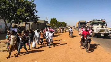 L'immagine mostra una scena di vita quotidiana lungo una strada in un ambiente urbano o semi-urbano. Si notano molte persone che camminano lungo il lato della strada, alcune con sacchi o beni in mano, e altre che sembrano discutere tra di loro. Sul lato opposto della strada ci sono veicoli in transito, tra cui camion e motociclette. La vegetazione circostante è presente, con alcuni alberi che offrono ombra. Il terreno appare polveroso, e l'atmosfera complessiva sembra vivace e movimentata.