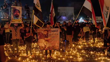 L'image montre une foule rassemblée dans une rue sombre, illuminée par des centaines de bougies. Les personnes portent des affiches et des banderoles, certaines mettant en avant des portraits ou des messages en hommage à des individus, probablement des victimes ou des figures importantes. Il y a aussi des drapeaux en arrière-plan, suggérant un contexte national. L'atmosphère semble être à la commémoration ou au rassemblement pour une cause, avec un sentiment de solidarité visible parmi les participants.