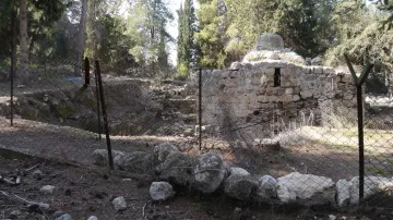 L'image montre un site archéologique entouré d'une clôture en fil de fer. Il y a des ruines anciennes, probablement d'une structure en pierre, avec des éléments végétaux environnants tels que des arbres et des buissons. Le sol est rempli de pierres et de débris, ce qui donne une impression d'abandon. L'atmosphère semble paisible, mais aussi légèrement mélancolique, témoignant d'un passé oublié.