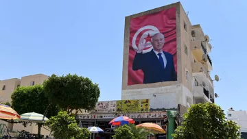 La imagen muestra un gran mural en la pared de un edificio, donde se puede ver a un hombre de pie y saludando. Detrás de él hay una bandera que parece ser de Túnez. En la parte inferior de la imagen, se observa un pequeño mercado o tienda al aire libre, con sombrillas de colores y algunas personas. Hay árboles y bicicletas en el entorno, lo que sugiere un ambiente urbano y concurrido. El cielo está despejado y soleado.