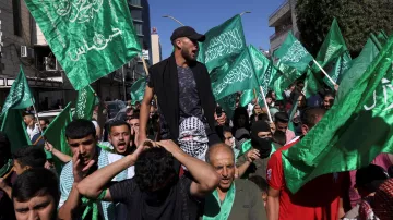 Cette image montre une manifestation animée, avec des personnes brandissant de grands drapeaux verts. On peut voir une foule de manifestants, certains sont en train de crier ou de chanter, tandis qu'un homme est porté sur les épaules de quelqu'un d'autre. L'ambiance semble énergique, et l'arrière-plan présente des bâtiments urbains.