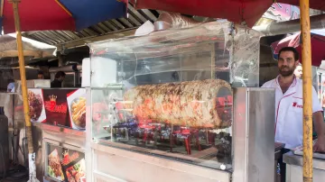 L'image montre un stand de nourriture de rue où un homme se tient derrière une vitrine en plastique transparente. À l'intérieur de la vitrine, il y a un grand morceau de viande en train de rôtir sur des braises, probablement un kebab ou un gyros. Le stand est couvert de parasols colorés, et il y a des affiches annonçant les plats proposés. L'ambiance semble vivante, typique des marchés animés de rues.