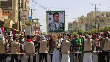 The image depicts a large crowd gathered for a rally or protest in a city, likely in Yemen. In the foreground, a group of people, some wearing beige vests, are standing behind a barricade. They are holding a large poster featuring a portrait of a man, who appears to be a political or religious leader, alongside images of other individuals. The crowd is holding flags, and buildings can be seen in the background, suggesting an urban setting. The atmosphere seems to convey a sense of solidarity or support for the figure on the poster.