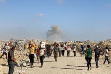 Des personnes marchent dans des décombres, portant des colis, avec de la fumée au loin.