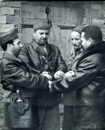 L'image montre quatre hommes serrant les mains les uns des autres dans une posture de camaraderie ou de négociation. Ils portent des tenues militaires ou de combat, et l'environnement semble être extérieur, avec des murs en pierre en arrière-plan. L'expression de leurs visages peut suggérer la détermination ou la concentration, et l'interaction semble exprimer un moment significatif.