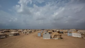 L'image montre un paysage désertique avec des tentes disposées dans une vaste étendue de sable. Le ciel est nuageux, avec des nuages gris et blancs qui donnent une ambiance tranquille mais assez chargé. Les tentes sont de couleurs variées, majoritairement beige et grise, et semblent être organisées de manière assez dense, suggérant qu'il s'agit d'un camp, probablement pour des personnes déplacées ou réfugiées. L'environnement général est aride et peu végétalisé, accentuant l'aspect isolé du lieu.