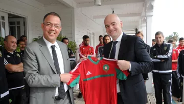 L'image montre deux hommes souriants qui se tiennent devant un groupe de personnes en arrière-plan. L'un d'eux porte un costume gris et tient un maillot de football rouge et vert, probablement aux couleurs de l'équipe nationale marocaine. L'autre homme, en costume sombre, semble également heureux et participe à la remise de ce maillot. Les autres personnes en arrière-plan portent des ensembles sportifs noirs avec des détails blancs. L'ambiance suggère un moment de célébration ou d'échange, probablement lié au football.