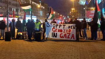 L'image montre un groupe de manifestants rassemblés dans une rue, tenant une grande banderole sur laquelle il est écrit "STOPPT DEN GENOZID IN GAZA". Ils portent des drapeaux palestiniens et semblent exprimer leur soutien à Gaza. L'atmosphère est sérieuse, et il est probable que la manifestation s'inscrive dans un contexte de protestation contre des violences ou des injustices en Palestine. Les lumières des bâtiments environnants suggèrent qu'il s'agit d'une manifestation en soirée.