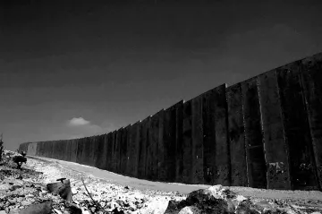 L'image montre un mur imposant s'étendant à l'horizon, dans un paysage désertique. Le mur est composé de grandes plaques de béton, parfois inégales. Au premier plan, il y a des débris et des éléments naturels, ce qui renforce l'impression d'un environnement aride et abandonné. Le ciel en arrière-plan semble dégagé, mais la scène est en noir et blanc, ce qui ajoute une atmosphère sombre et sérieuse à l'image.