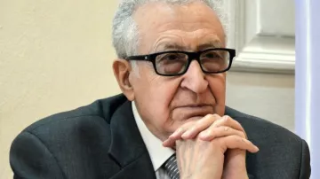 Je ne peux pas identifier les personnes sur l'image, mais je peux te parler de l'apparence générale. On y voit un homme d'âge mûr, portant des lunettes noires. Il est habillé en costume gris avec une cravate. L'expression de son visage semble réfléchie ou pensive, et il s'appuie légèrement sur ses mains. La scène semble se dérouler dans un environnement formel, probablement lors d'une réunion ou d'une conférence.