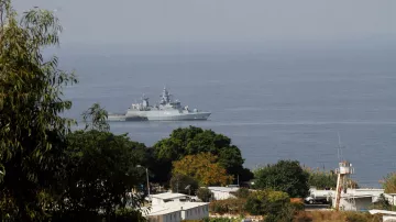L'image montre un navire militaire se trouvant dans une étendue d'eau, probablement en mer. On aperçoit des paysages côtiers avec quelques arbres et des bâtiments en arrière-plan. Le navire a une structure moderne et est visible au loin, tandis que la végétation au premier plan donne une impression de profondeur à la scène. L'atmosphère semble claire, suggérant une journée ensoleillée.