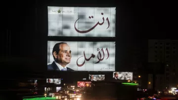 The image shows large billboards prominently displaying Arabic text. One of the billboards features a portrait of a man, likely a political figure, along with the words "انت الأمل," which translates to "You are the hope." The setting appears to be a busy urban area at night, with car lights creating a dynamic effect, suggesting movement and activity. The overall atmosphere reflects a lively city scene with political messaging.