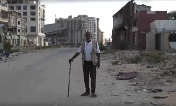 Un homme âgé avec une canne se tient sur une rue déserte, entouré de bâtiments en ruine.