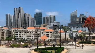 صورة لمدينة حديثة، مباني مرتفعة، سماء زرقاء وأشجار. يبدو النشاط العمراني واضحًا.