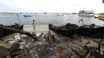 L'image montre une scène côtière après un incendie. On peut y voir des bateaux endommagés et carbonisés sur le rivage. Un homme se tient près de l'eau, regardant vers le horizon. La mer semble calme, avec plusieurs petites embarcations au loin. Le ciel est nuageux, et le paysage est marqué par les conséquences de l'incendie, avec des débris et des cendres sur le sable.