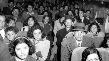 L'image montre l'intérieur d'un avion rempli de passagers. On peut voir un groupe diversifié de personnes, y compris des adultes et des enfants, assis sur des rangées de sièges. Les expressions des passagers varient, certains semblent sérieux, tandis que d'autres regardent droit devant. L'ambiance semble être celle d'un voyage, probablement dans un contexte historique. L'éclairage de l'image est en noir et blanc, ce qui lui donne une atmosphère vintage.