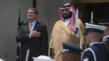 L'image montre deux hommes en tenue formelle. L'un est sur le devant, ayant la main sur le cœur, tandis que l'autre se tient à ses côtés. Ils sont entourés de membres d'une garde d'honneur, portant des uniformes militaires. L'environnement semble officiel, probablement un événement diplomatique ou militaire. Les deux hommes ont l'air sérieux et engagés dans la cérémonie.