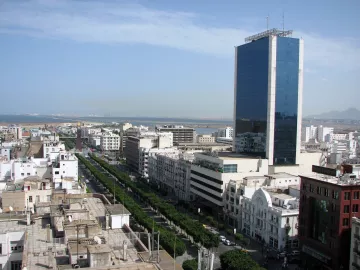 L'image montre un paysage urbain, probablement d'une ville côtière avec une vue panoramique. On peut voir de grands bâtiments modernes, dont une tour en verre qui se dresse au centre. La rue est bordée d'arbres et d'immeubles, créant une ambiance sympathique. À l'arrière-plan, on aperçoit l'océan, suggérant que la ville est située près de la mer. Le ciel est dégagé avec quelques nuages, indiquant une belle journée.